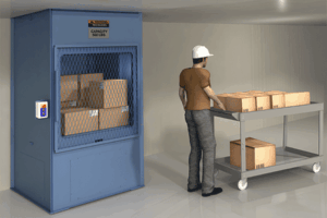 A man wearing a white hard hat stands beside a cart with boxes, facing a large blue industrial lift with a mesh door labeled “CAPACITY 500 LBS.” in a warehouse setting.