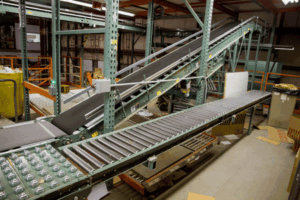 A conveyor system with metal rollers and an inclined belt transports packages in a warehouse with industrial shelving and scattered boxes.