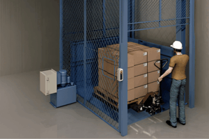 A worker wearing a hard hat moves a pallet jack loaded with stacked boxes into a metal cargo lift enclosed by a wire mesh cage in an industrial setting.