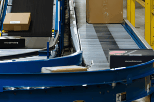Conveyor belts transport cardboard boxes and packages through a warehouse or distribution center, with blue guardrails and yellow support structures visible.