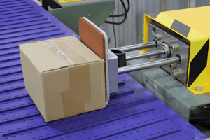A cardboard box is pushed along a purple conveyor belt by a mechanical arm from yellow automated packaging equipment.