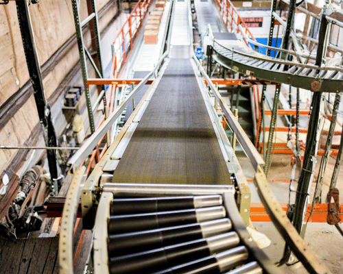 A conveyor belt system in a warehouse, with empty rollers in the foreground and shelves of boxes and packages visible in the background.