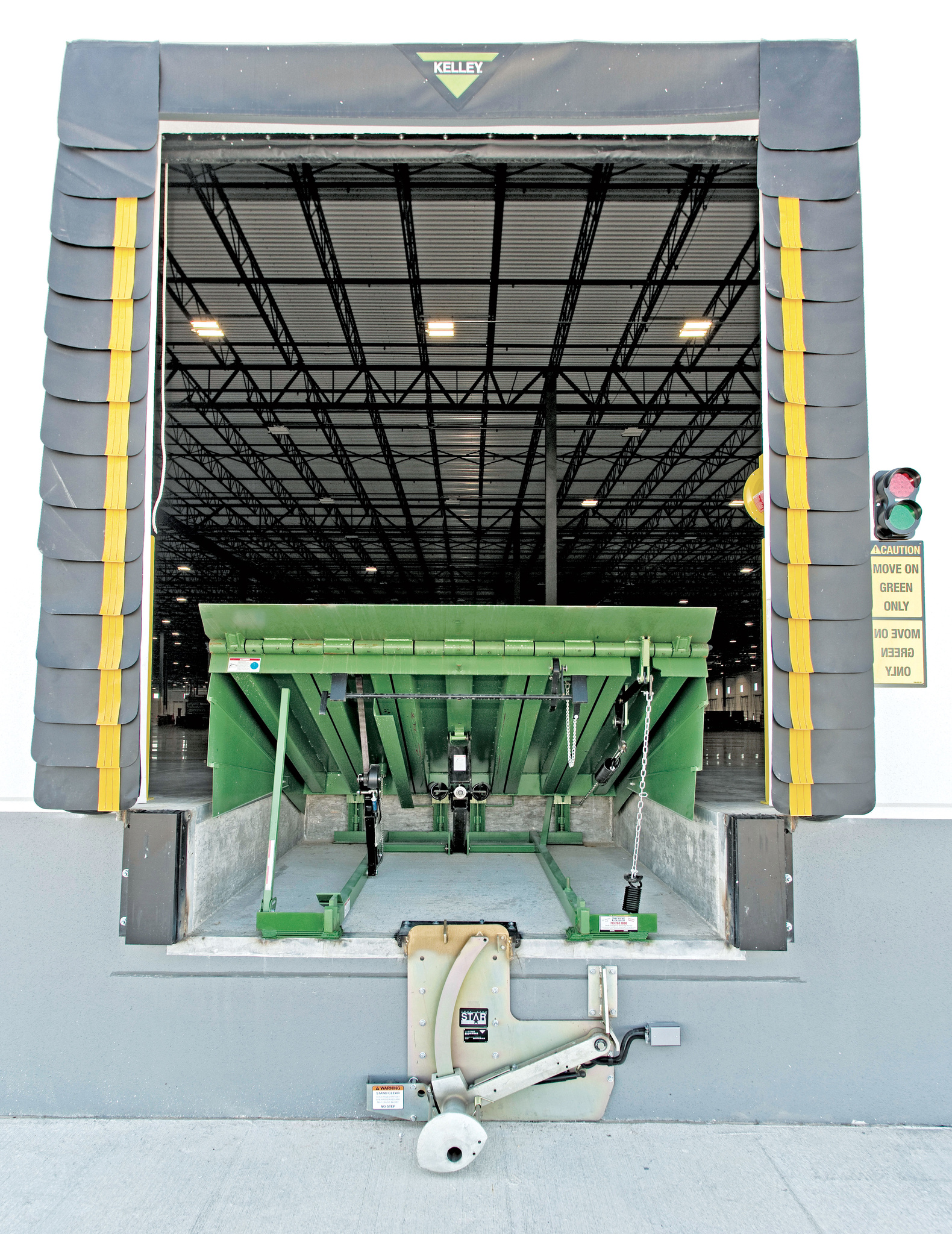 A green loading dock leveler is raised at an open industrial warehouse door, with a padded frame and a move on green only safety sign visible on the right. The large warehouse interior appears empty.