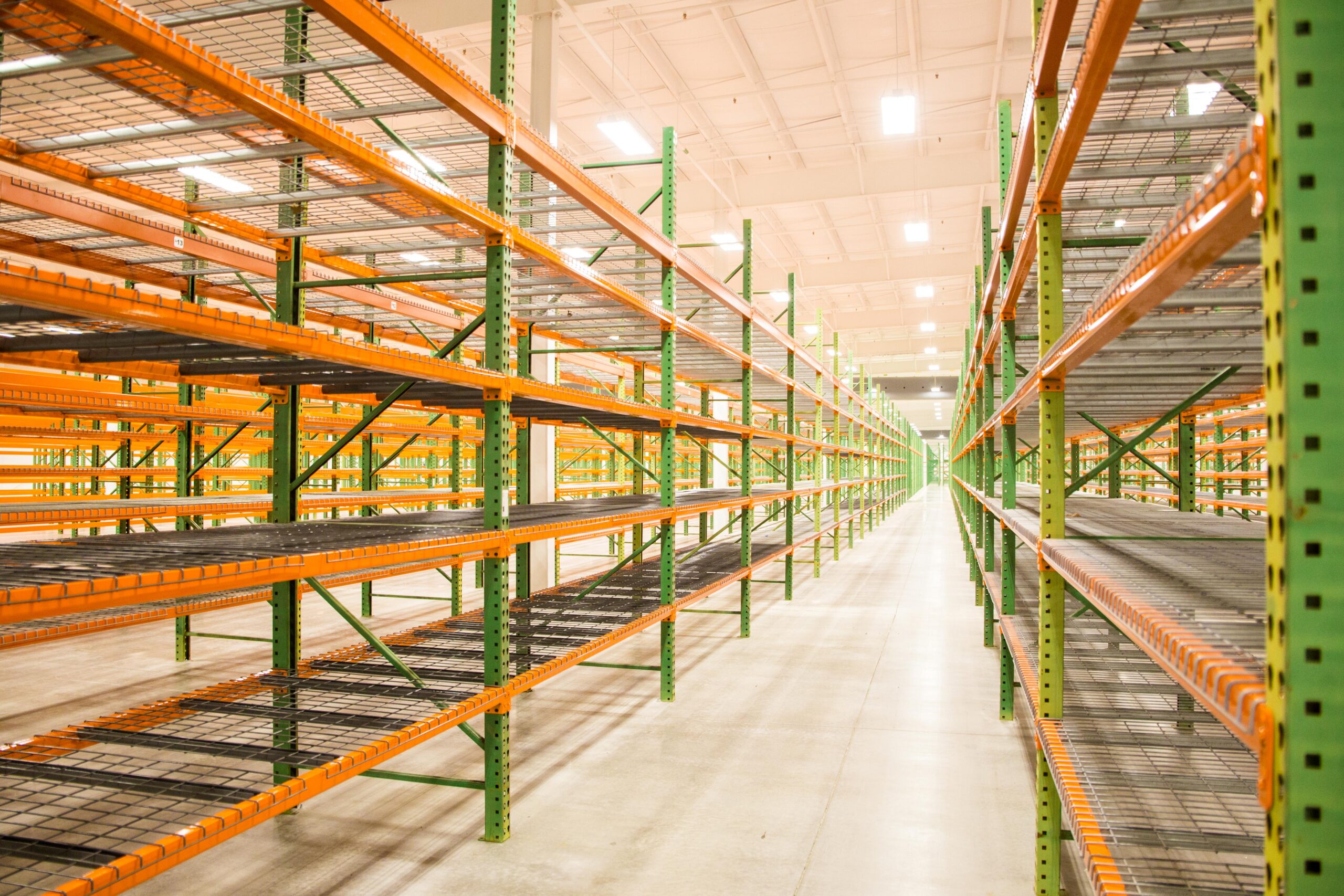 Rows of empty green and orange metal shelves stretch down a brightly lit warehouse with a smooth concrete floor and high ceiling.