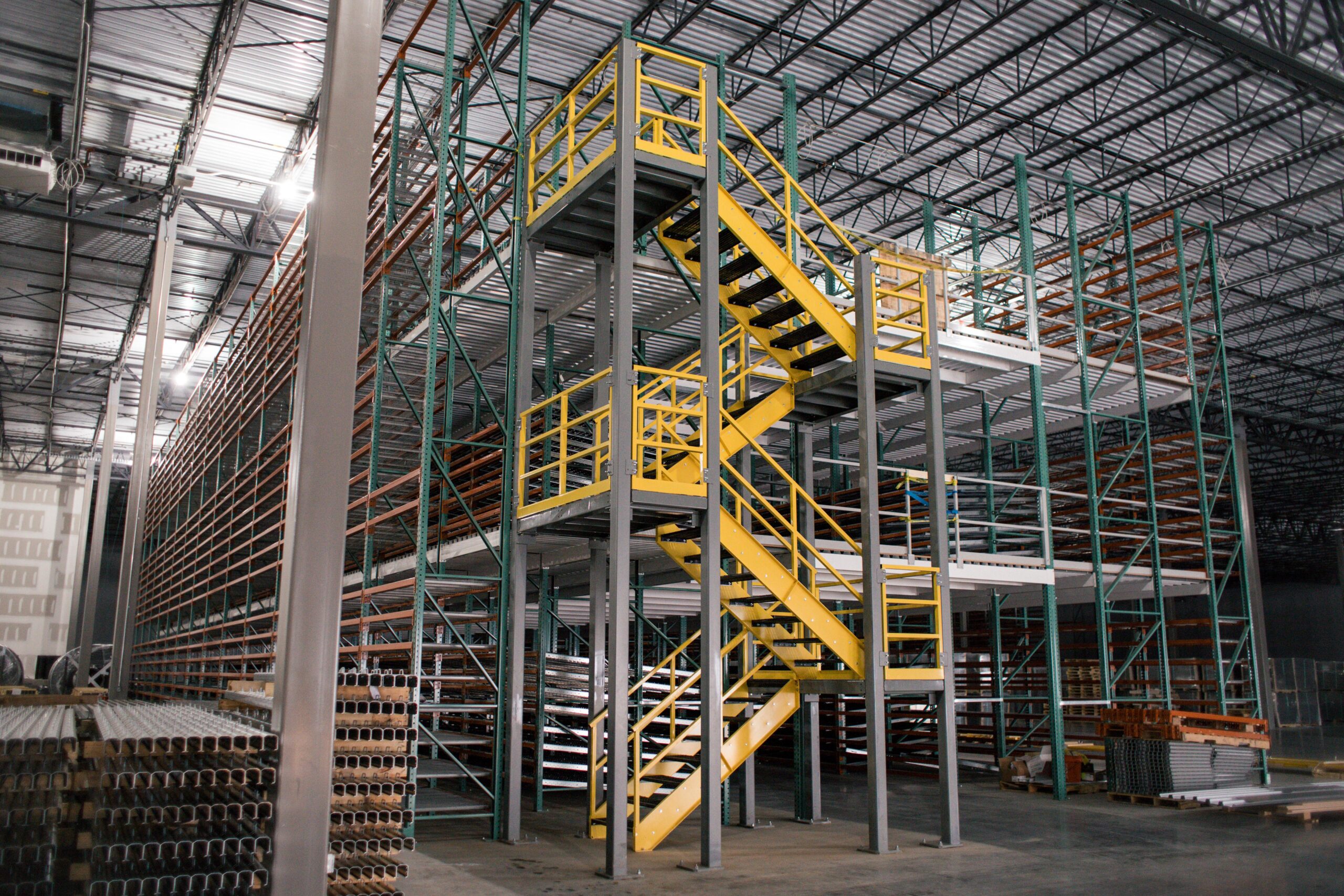 Metal warehouse interior with multi-level industrial shelving and a yellow staircase in the center. Various metal pipes and materials are stacked on shelves and on the floor. The ceiling is high with exposed beams and lighting.