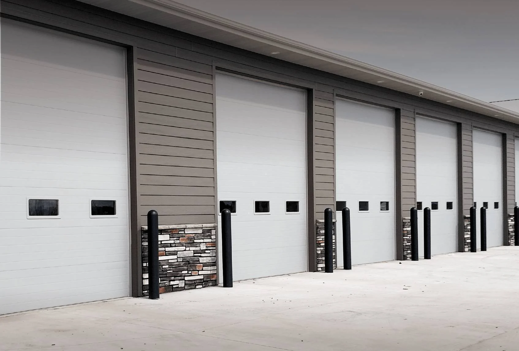 Five large, white industrial garage doors are lined up on a modern building with gray siding and stone accents at the base. Black bollards are positioned in front of each door for protection.