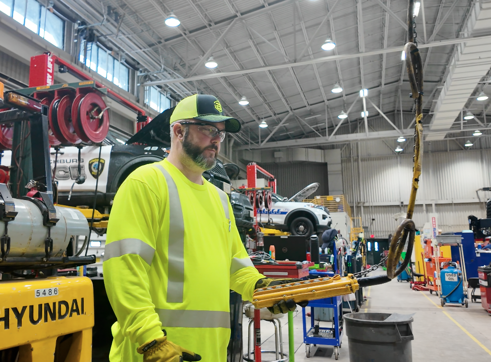 A man in a neon yellow safety shirt and cap operates lifting equipment in a busy automotive repair shop with cars and tools in the background.
