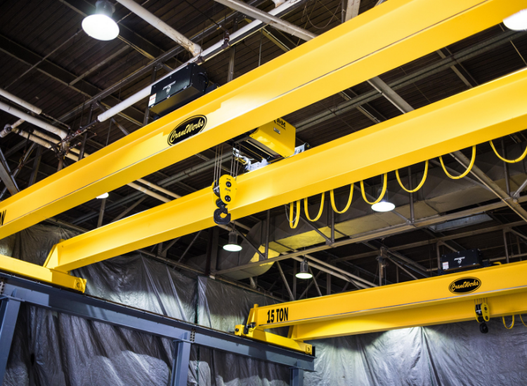 Three large yellow overhead cranes with 15 TON labels are installed in an industrial facility, with metal beams and gray tarps in the background. The ceiling and lights of the warehouse are visible above.